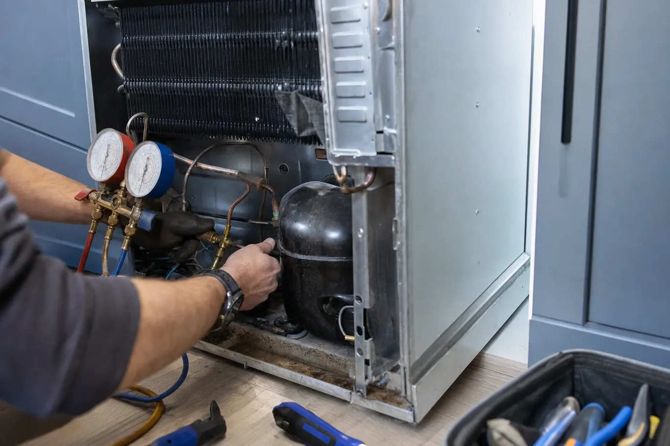 A hand holding a two-gauge manifold set at the open lower bay of a built-in refrigerator, the finned condenser coil above and the compressor dome below