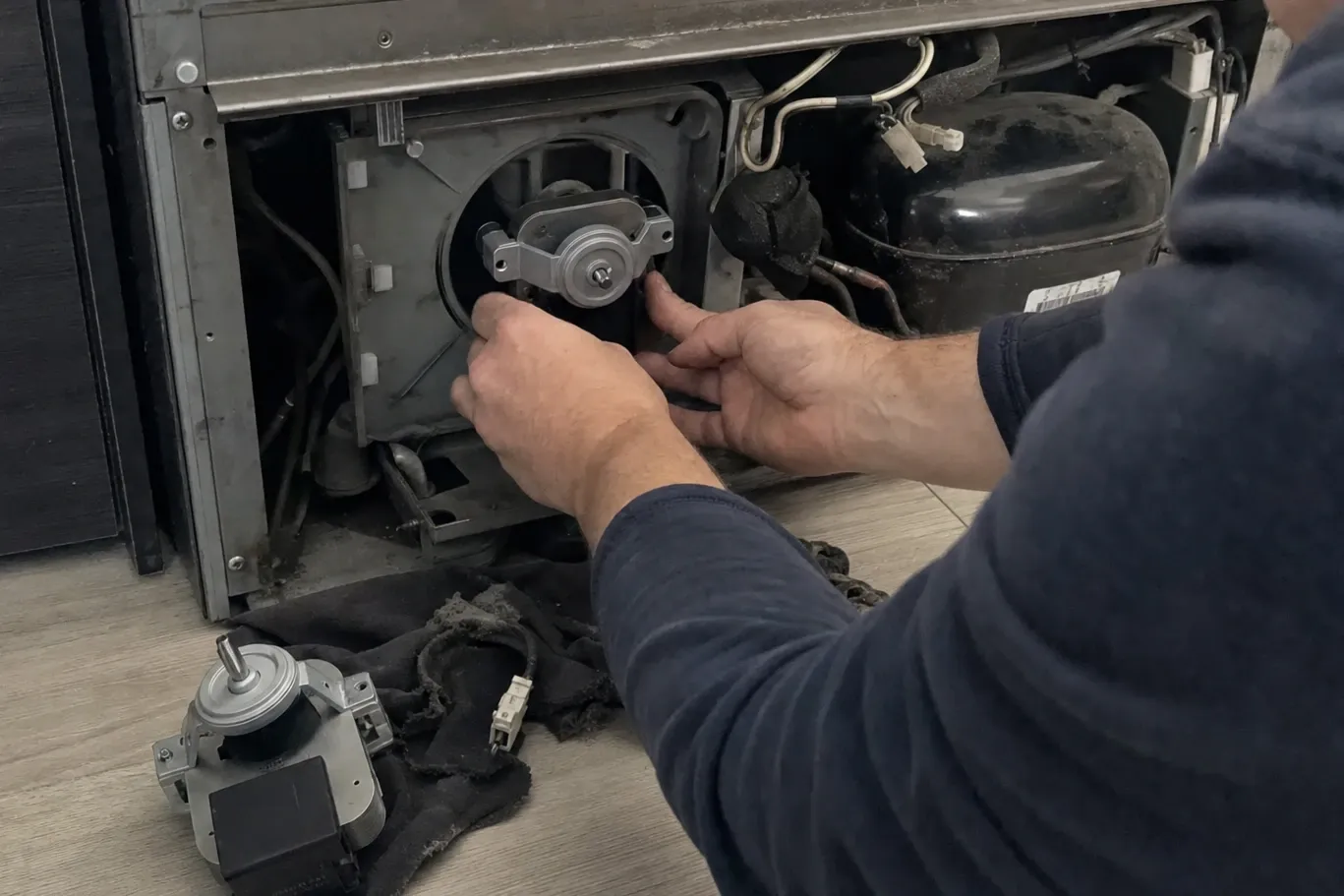 Two hands seating a replacement fan motor into its bracket inside the machine compartment of a refrigerator, the old motor lying on a cloth on the floor